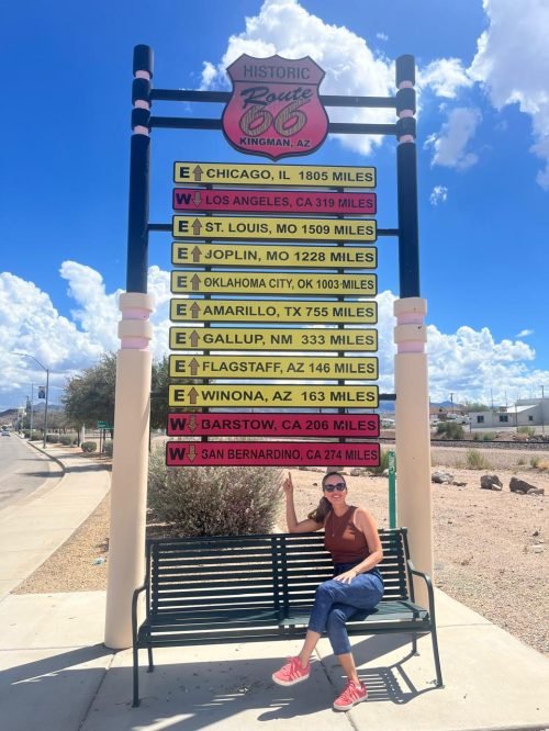 Luciana sentada no banco com a placa das cidades da Rota 66 de Chicago a San Bernardino, em Kingman, AZ.